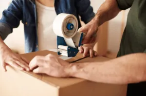 Un pacco che viene chiuso da un uomo con del nastro bianco mentre una donna lo aiuta Un pacco che viene chiuso da un uomo con del nastro bianco mentre una donna lo aiuta