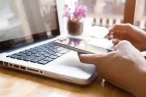 Persona che tiene in mano il suo cellulare mentre controlla il computer Persona che tiene in mano il suo cellulare mentre controlla il computer